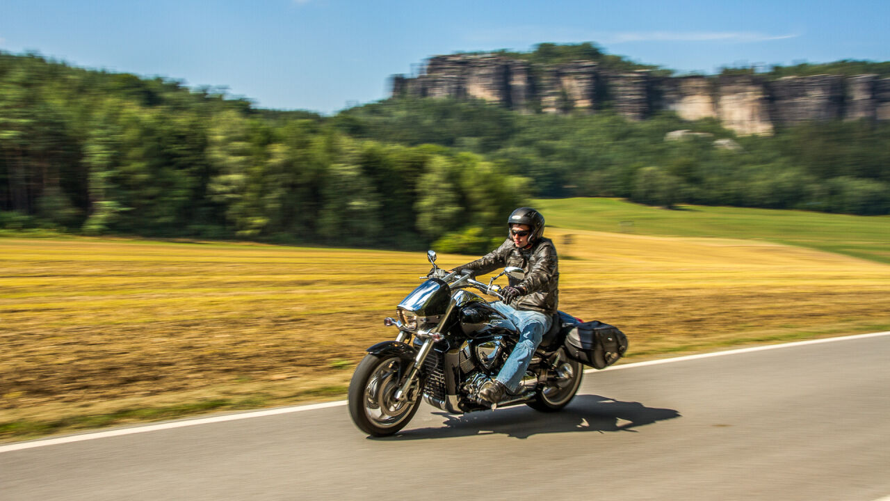 Motorradfahrer auf einer Landstraße vor Getreidefeld, dahinter Sandsteinfelsen über dem Wald Motorradfahrer auf einer Landstraße vor Getreidefeld, dahinter Sandsteinfelsen über dem Wald